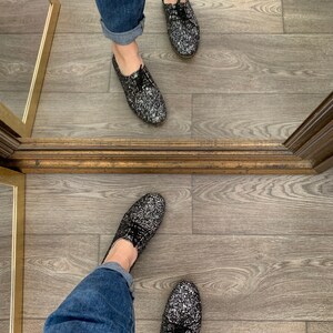 May include: A pair of black glitter shoes with black laces. The shoes are worn by a person wearing blue jeans.