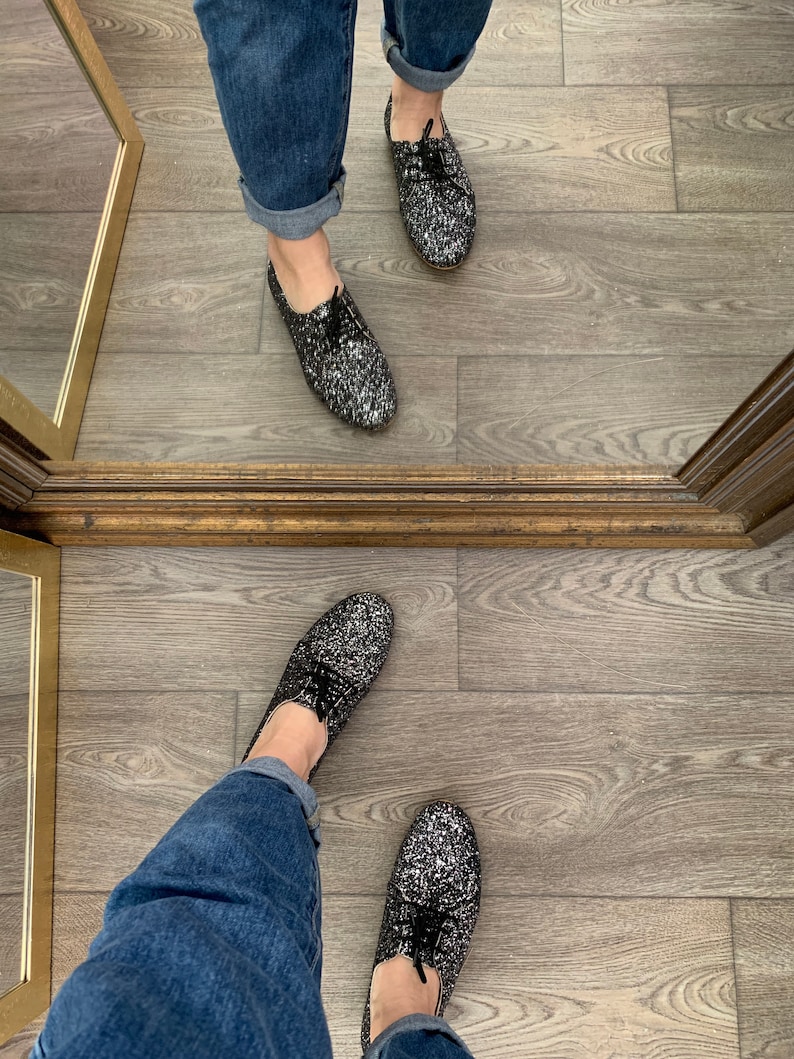 May include: A pair of black glitter shoes with black laces. The shoes are worn by a person wearing blue jeans.