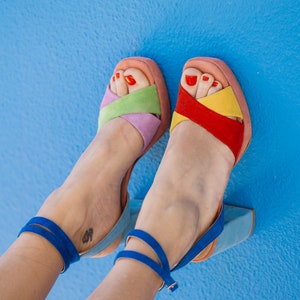 May include: A pair of colorful platform sandals with a blue ankle strap. The sandals feature a mix of pastel colors, including pink, green, purple, yellow, and red. The background is a solid blue.