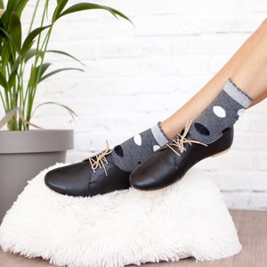 May include: A pair of black leather lace-up shoes with a low heel. The shoes are resting on a white fluffy rug. The shoes are worn with gray and white polka dot socks.