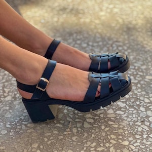 May include: Black leather sandals with a chunky heel and a woven strap design. The sandals have a buckle closure and a thick sole. The shoes are shown on a textured surface.