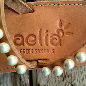 May include: Close-up of a pair of tan leather sandals with white pearl embellishments. The sandals have the brand name "aelia GREEK SANDALS" embossed on the leather.