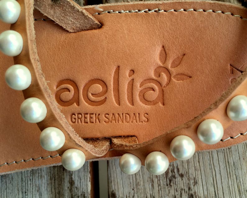 May include: Close-up of a pair of tan leather sandals with white pearl embellishments. The sandals have the brand name "aelia GREEK SANDALS" embossed on the leather.