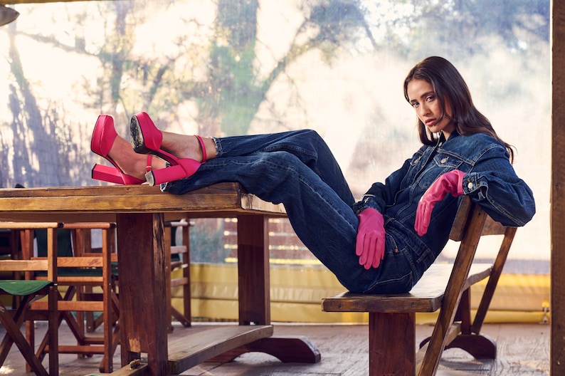 Puede incluir: Una mujer con una chaqueta vaquera y pantalones vaqueros se sienta en un banco de madera con las piernas apoyadas en una mesa de madera. Lleva tacones de plataforma rosa y guantes rosas.
