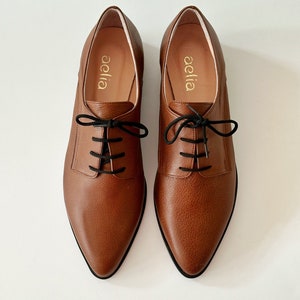 May include: A pair of brown leather lace-up shoes with a pointed toe. The shoes have black laces and the brand name "aella" is printed on the inside of each shoe.