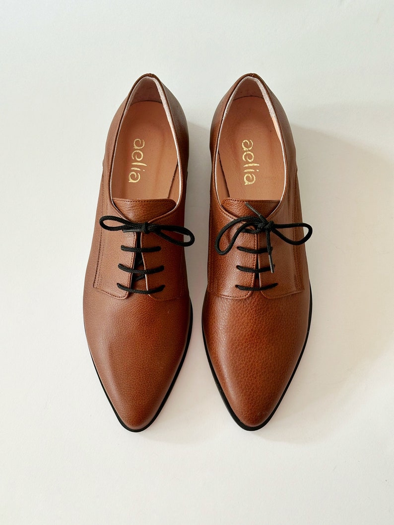 May include: A pair of brown leather lace-up shoes with a pointed toe. The shoes have black laces and the brand name "aella" is printed on the inside of each shoe.