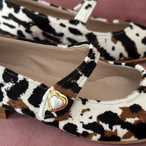 May include: A pair of white, black, and brown cow print ballet flats with a gold heart-shaped buckle. The buckle has a white pearl center.