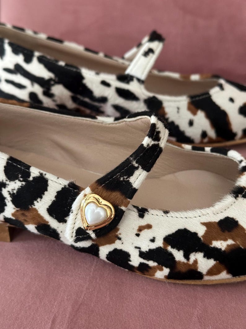 May include: A pair of white, black, and brown cow print ballet flats with a gold heart-shaped buckle. The buckle has a white pearl center.