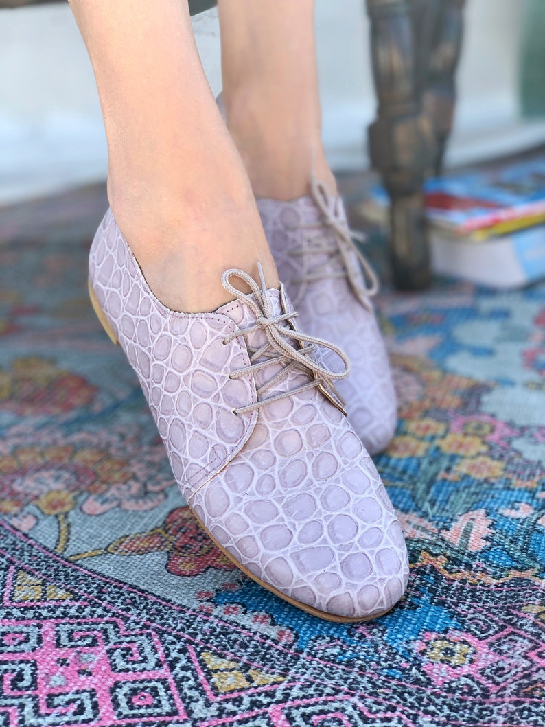 Pu&ograve; includere: Un paio di oxford in pelle color lilla chiaro con un motivo goffrato a coccodrilo. Le scarpe hanno i lacci e sono appoggiate su un tappeto colorato.