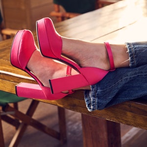 Puede incluir: Un par de zapatos de tac&oacute;n alto de color rosa brillante con correas de tobillo. Los zapatos est&aacute;n sobre una mesa de madera con un par de sillas de madera en el fondo.