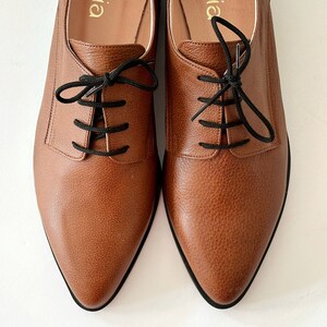 May include: A pair of brown leather oxfords with black laces. The shoes have a pointed toe and a low heel. The brand name "aellia" is printed on the inside of each shoe.