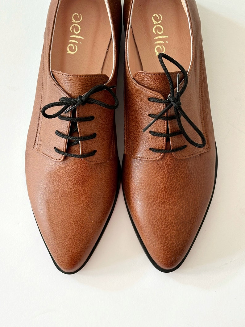 May include: A pair of brown leather oxfords with black laces. The shoes have a pointed toe and a low heel. The brand name "aellia" is printed on the inside of each shoe.