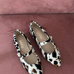 May include: A pair of white and brown leopard print ballet flats with a gold buckle and the word "aelia" written on the side.
