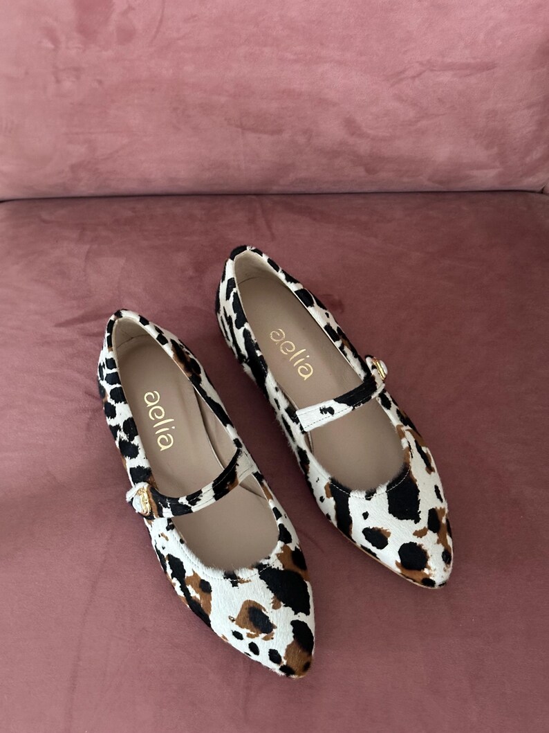 May include: A pair of white and brown leopard print ballet flats with a gold buckle and the word "aelia" written on the side.