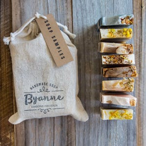 May include: A beige drawstring fabric pouch with a brown paper tag labelled SOAP SAMPLES and the text Byanne Handmade Soap Canberra Australia, accompanied by a vertical stack of eight rectangular soap bars featuring various botanical inclusions.