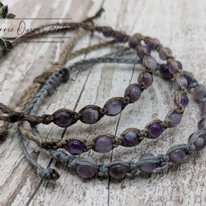 May include: Three macrame bracelets with amethyst beads. The bracelets are in shades of brown and gray, with the amethyst beads adding a pop of purple. The bracelets are displayed on a wooden surface. The text "Kerrie Owens Arts" is visible.