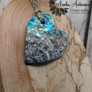 May include: A heart-shaped pendant crafted from iridescent abalone shell, showing blues, greens, and silver. It hangs from a natural fibre cord. The image also includes the text "Collection Aroha Aotearoa. Ocean edition".
