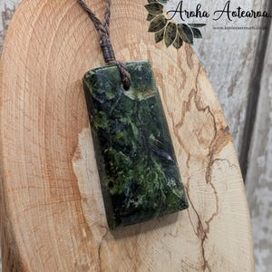 May include: A rectangular green stone pendant with a dark brown cord. The stone has a marbled appearance with dark green and black swirls. The pendant is part of the "Aroha Aotearoa" collection.