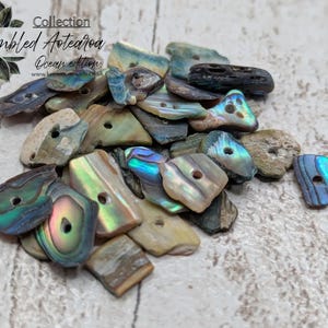 May include: A collection of tumbled Aotearoa Ocean edition buttons in various shapes and sizes. The buttons are made from iridescent abalone shells, displaying shades of blue, green, and purple. Some buttons have a natural, weathered appearance, while others have a polished, glossy finish.