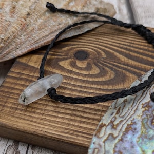 May include: A black braided cord bracelet with a clear quartz crystal pendant. The bracelet is lying on a wooden surface with a shell in the background.