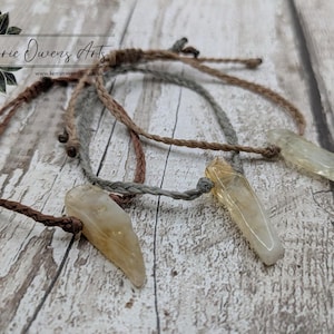 May include: Three necklaces with light brown, grey, and dark brown braided cords. Each necklace features a clear, elongated crystal pendant with a yellow tint. The background is a light wood surface. The text "Kerrie Owens Arts" is visible.