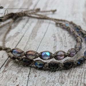 May include: Two macrame bracelets with iridescent beads. The beads are in shades of purple, blue, and green. The bracelets are made of a natural-colored cord and are displayed on a wooden surface. The bracelets are handmade.