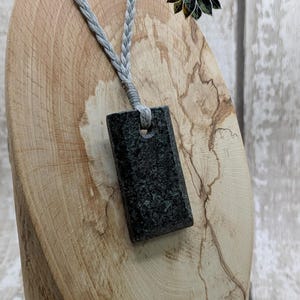 May include: A dark gray stone pendant with a hole in the top, hanging from a gray braided cord. The pendant is on a wooden display stand. The text "Collection Ancestral fusión. www.kerrieowensarts.co.uk" is visible in the top right corner of the image.