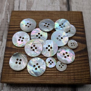 May include: A collection of iridescent white mother of pearl buttons with four holes each. The buttons are arranged in a random pattern on a wooden surface.