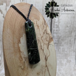 May include: A green jade pendant with a black cord necklace. The pendant is carved in a rectangular shape with rounded edges. The pendant is on a wooden surface.