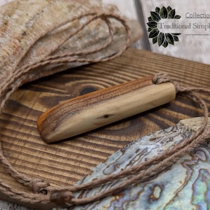 May include: A wooden pendant with a natural wood finish hangs from a brown braided cord. The pendant is a simple, rectangular shape with rounded edges. The cord is tied with a knot at the top of the pendant. The pendant is displayed on a wooden surface with a natural wood grain.