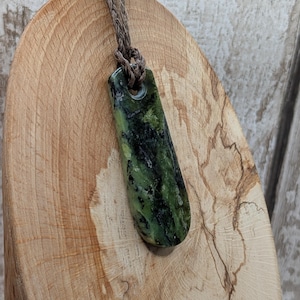 May include: A green and black stone pendant with a braided brown cord. The rectangular pendant has a hole at the top for the cord. The stone has a marbled appearance with dark green and black patterns. The pendant is displayed on a wooden surface.