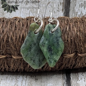 May include: A pair of green stone earrings with silver-colored hooks. The earrings are a natural, irregular shape and have dark green and black marbling. The earrings are displayed on a woven brown surface. The text "Collection Tumbled Aotearoa" is visible in the background.