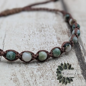May include: A close-up of a handmade bracelet featuring green and white gemstone beads. The beads are strung on a brown, woven cord. The bracelet is displayed on a light-colored, textured surface. The overall aesthetic is natural and earthy.