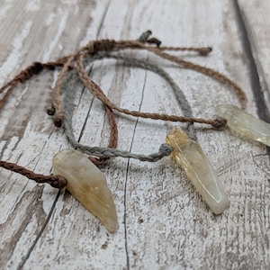 May include: Three bracelets with brown, grey, and beige cords and a light brown, pointed crystal charm on each.