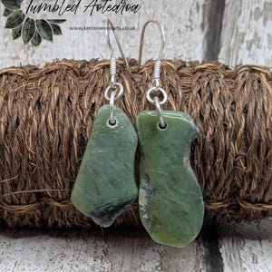 May include: A pair of green stone earrings with silver-colored hooks. The earrings are irregularly shaped and have a polished surface. The background includes the text "Collection Tumbled Aotearoa" and a website address.