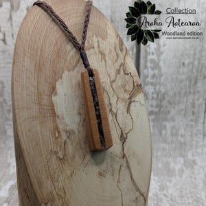 May include: A wooden pendant necklace with a brown cord. The pendant is made of wood with a dark brown stripe down the center. The pendant is hanging on a wooden display stand. The text "Aroha Aotearoa Woodland edition" is visible in the upper right corner of the image.