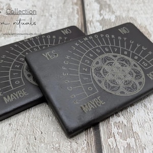 May include: Two dark gray planchette boards with engraved designs. Each board features a circular design, letters, numbers, and the words "YES", "NO", and "MAYBE". The boards are part of the "Modern rituals" collection.