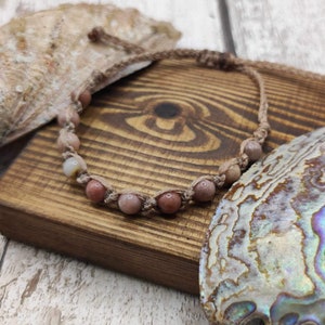 May include: A pink and white beaded bracelet with a brown cord. The bracelet is on a wooden surface with a seashell.