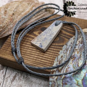 May include: A gray braided cord necklace with a light blue and white ceramic pendant. The pendant is a rectangular shape with rounded edges. The necklace is displayed on a wooden surface with a seashell.