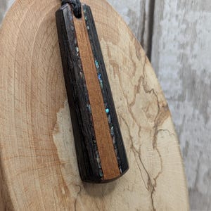 May include: A rectangular pendant featuring a central strip of light brown wood flanked by two dark brown wood sections. Inlaid between the wood pieces are thin strips of iridescent abalone shell fragments. The pendant hangs from a dark gray cord.