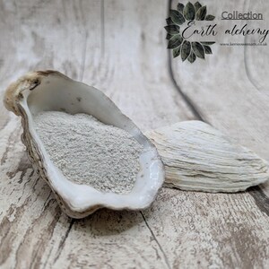 May include: An oyster shell filled with pale grey powder, alongside a shell fragment, on a light wood-effect surface. The image includes the text "Collection Earth alchemy." and the website address.