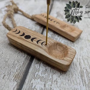 May include: Two rectangular light brown wooden incense holders featuring a burned-in moon phase design and a circular indentation. Each holder has a thin metal rod inserted for incense and a thick jute rope loop attached to one end for hanging.