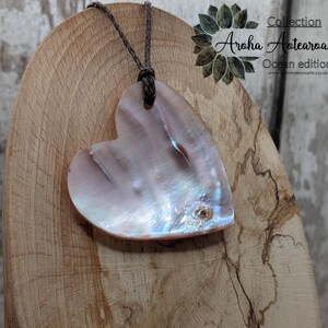 May include: A heart-shaped pendant crafted from iridescent mother-of-pearl, suspended from a brown cord. The pendant displays a pearlescent sheen with pink and blue hues. The text "Aroha Aotearoa. Ocean edition" is visible.