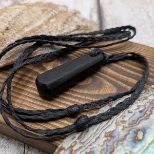 May include: A black rectangular pendant with a smooth, matte finish suspended from a thin, braided black cord. The pendant features a single hole at the top for the cord to pass through, creating a simple, linear design with a dark, uniform colour.