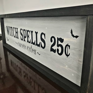 Puede incluir: Un letrero enmarcado en negro con pintura blanca y letras negras que dice "Witch Spells 25&cent; ~curses extra~" con una peque&ntilde;a silueta de murci&eacute;lago negro en la esquina superior derecha.