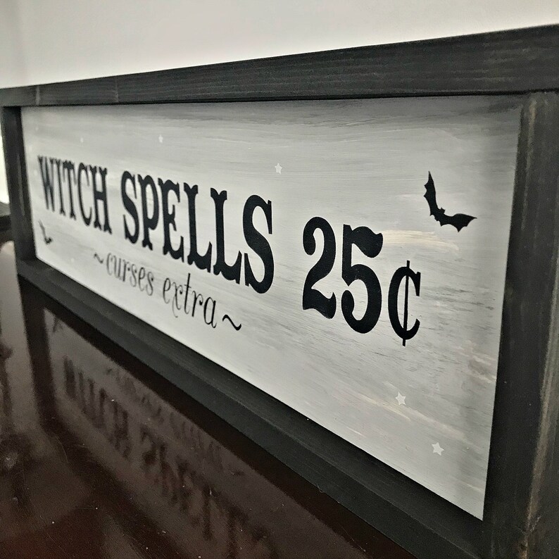 Puede incluir: Un letrero enmarcado en negro con pintura blanca y letras negras que dice "Witch Spells 25&cent; ~curses extra~" con una peque&ntilde;a silueta de murci&eacute;lago negro en la esquina superior derecha.