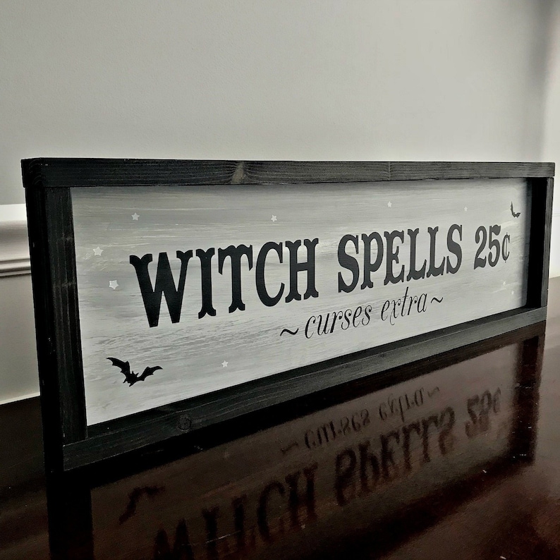 Puede incluir: Un letrero de madera enmarcado en negro con fondo blanco y letras negras que dice "Witch Spells 25&cent; ~ curses extra ~". El letrero tiene una peque&ntilde;a silueta de murci&eacute;lago negro en la esquina inferior izquierda.