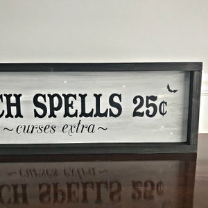 Puede incluir: Un letrero en blanco y negro con fondo gris que dice "Witch Spells 25&cent; ~ curses extra ~". El letrero est&aacute; enmarcado en negro y tiene una peque&ntilde;a silueta de murci&eacute;lago en la esquina superior derecha.