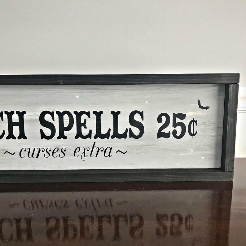 Puede incluir: Un letrero en blanco y negro con fondo gris que dice "Witch Spells 25&cent; ~ curses extra ~". El letrero est&aacute; enmarcado en negro y tiene una peque&ntilde;a silueta de murci&eacute;lago en la esquina superior derecha.