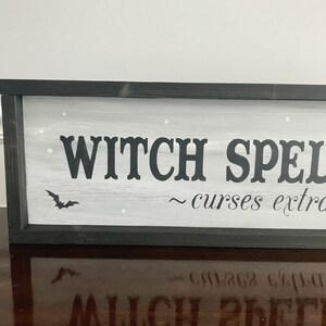 Puede incluir: Un letrero de madera enmarcado en negro con fondo blanco y letras negras que dice "WITCH SPELLS ~ curses extra". El letrero tiene una peque&ntilde;a silueta de murci&eacute;lago negro y estrellas blancas pintadas.